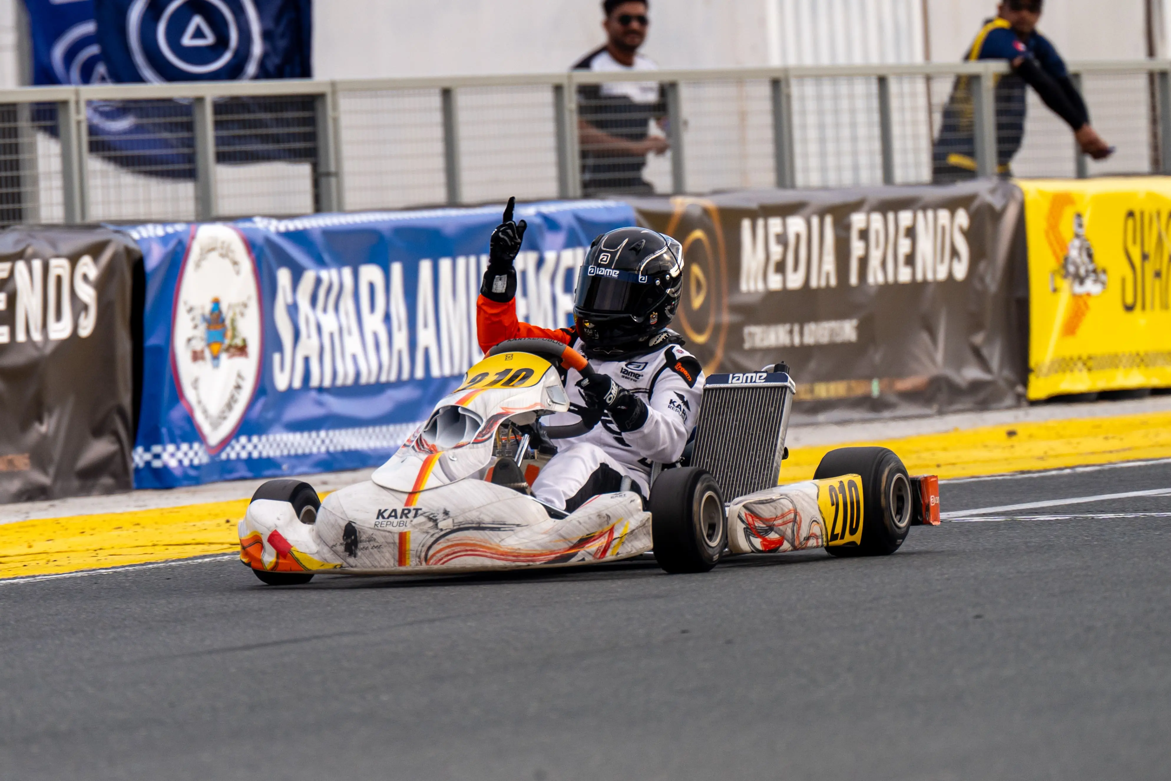 A young kart driver celebrates a win, arm raised, past the Media Friends trackside banner