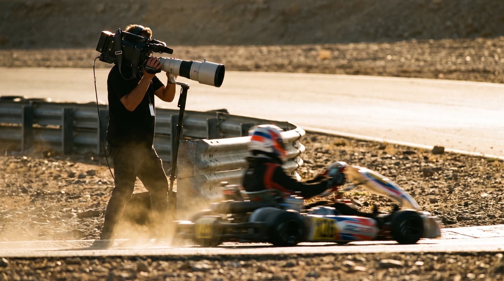 A camera operator with a broadcast camera tracking a single kart from the trackside barrier